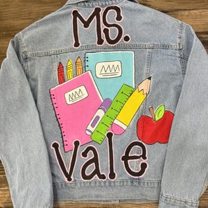 May include: Light blue denim jacket with hand-painted school-themed designs. Features the text "MS. Vale" in black lettering, along with colorful illustrations of notebooks, pencils, a crayon, a ruler, and an apple.
