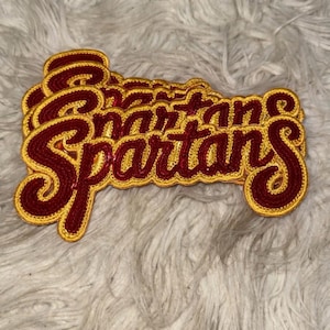 Puede incluir: Un conjunto de parches bordados con la palabra "Spartans" en una fuente cursiva. El texto es de color rojo oscuro, delineado con hilo dorado. Los parches son probablemente para ropa o accesorios.