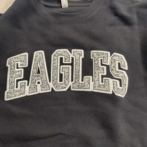 May include: Black crewneck sweatshirt with the word "EAGLES" in large, white, rhinestone-studded letters across the chest. The sweatshirt appears to be made of a soft material.