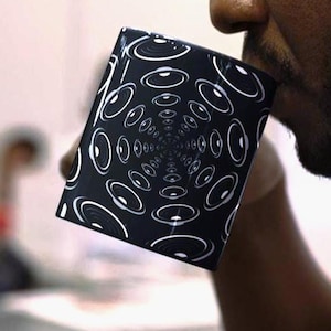 May include: A black and white coffee mug with a repeating concentric circle design. The mug is held up to a person's mouth, suggesting they are drinking from it. The design creates a visual tunnel effect.