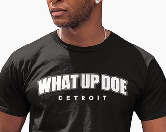 May include: A black short-sleeved crew neck t-shirt featuring the white text WHAT UP DOE in a bold, arched font, with the word DETROIT printed in smaller white capital letters directly underneath the main phrase on the chest area.