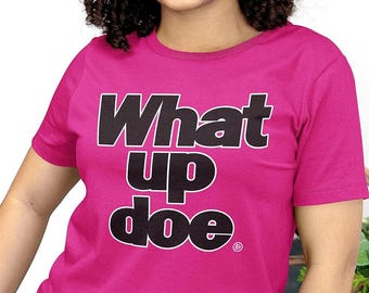 May include: A vibrant hot pink short-sleeved t-shirt featuring the text "What up doe." printed in bold black letters with a white outline across the chest. The shirt has a classic crew neckline and a standard fit, showcasing clear, legible typography.
