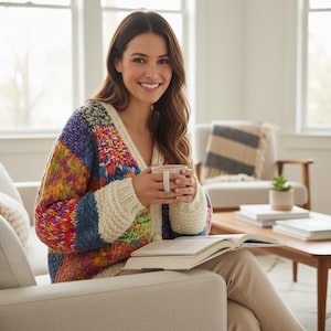 Può includere: Una donna indossa un cardigan lavorato a maglia grosso e colorato con scollo a V, tiene in mano una tazza e siede su un divano beige. Il cardigan presenta un design patchwork con vari colori. Sta leggendo un libro.