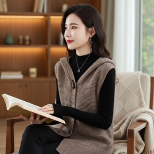 May include: A woman wearing a brown hooded vest over a black turtleneck and black pants. The vest has a button closure and a textured knit pattern. She is seated in a wooden chair and holding a book.