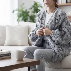 May include: A chunky knit gray cardigan with a cable knit design and bobble details. The open-front sweater has long sleeves and is worn over a white top. The person is sitting on a light-colored couch.