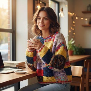 Pode incluir: Uma mulher usando um suéter colorido de retalhos, segurando uma caneca branca. O suéter apresenta blocos amarelos, azuis, vermelhos e roxos. Ela está sentada em uma mesa de madeira com um laptop e livros, perto de uma janela. A cena é acolhedora.