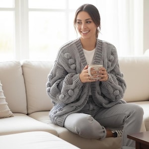 May include: A woman wearing a gray, chunky knit cardigan with large bobbles on the sleeves. The cardigan is open-front and paired with light-wash jeans. She is holding a white mug while sitting on a beige sofa.