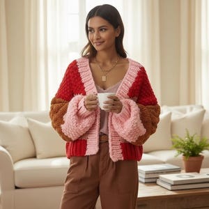 May include: A chunky, open-front cardigan in a color-block design. The sweater features red, pink, and brown sections. The sleeves are a voluminous, balloon style. The model is wearing the cardigan with brown pants and a gold necklace.