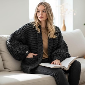 Puede incluir: Un cárdigan de punto grueso gris oscuro con mangas grandes y bolsillos. El suéter abierto lo lleva una persona sentada en un sofá de color claro, sosteniendo un libro. El cárdigan tiene un ajuste relajado y una apariencia acogedora.