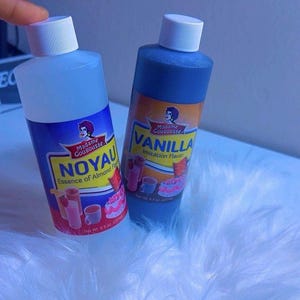 May include: Two bottles of Madame Gougousse flavoring extracts. One is labeled "NOYAU Essence of Almond Flavor" and the other "VANILLA Imitation Flavor". Both bottles are blue with white caps and colorful labels. The net weight is 8 fl oz (237 ml).