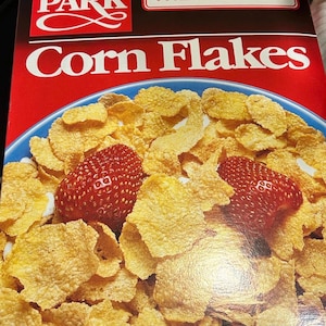 May include: A box of Hyde Park Corn Flakes with a serving suggestion. The box is red with the words "Corn Flakes" in large white letters. A bowl of corn flakes with two strawberries is shown.