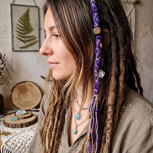 Puede incluir: Una mujer con largas rastas marrones, algunas adornadas con una envoltura morada, cuentas de madera y detalles decorativos. Lleva collares superpuestos con colgantes turquesas. La imagen tiene una estética bohemia.