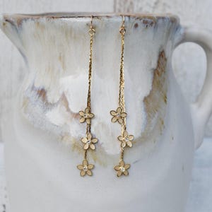 May include: Gold-toned dangle earrings featuring a delicate floral design. Each earring has a chain with three flower charms, each with a small, sparkling center. The earrings hang from a simple hook, showcasing a classic and elegant style.