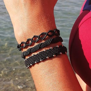 Könnte beinhalten: Drei schwarze Makramee-Armbänder werden an einem Arm gezeigt. Die Armbänder haben unterschiedliche Designs, darunter ein geflochtenes Muster und eines mit kleinen silbernen Perlen. Der Hintergrund zeigt Wasser und ein rotes Kleidungsstück.
