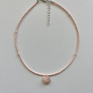 Peut inclure: Un collier délicat composé d'un fil de petites perles rose clair. Un petit pendentif en forme de coquillage rond est suspendu au centre. Le collier est doté d'un fermoir et d'une chaîne d'extension argentés. Le design est simple et élégant.