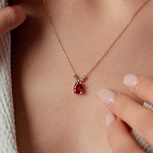 May include: A gold necklace with a small, teardrop-shaped, red gemstone pendant. The delicate chain is visible against the skin, and the pendant is the focal point. The necklace is a piece of jewelry.