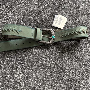 May include: A teal-colored belt with a decorative silver buckle and a small turquoise stone. The belt features stitched detailing along the edges. A white tag is attached to the belt.
