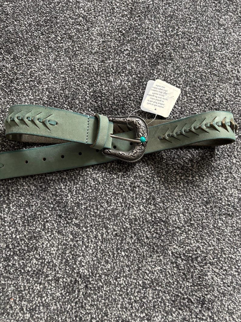 May include: A teal-colored belt with a decorative silver buckle and a small turquoise stone. The belt features stitched detailing along the edges. A white tag is attached to the belt.