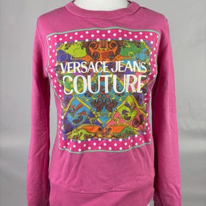 Puede incluir: Una sudadera rosa brillante con mangas largas. La parte delantera presenta un gráfico colorido con el texto "VERSACE JEANS COUTURE" en blanco. El gráfico está enmarcado por un borde de lunares blancos.