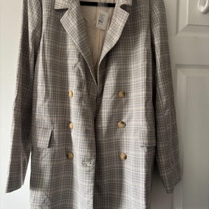 May include: A double-breasted blazer in a plaid pattern with tan, white, and gray tones. The blazer has a notched collar, two front pockets, and four buttons. The brand name "FREE PEOPLE" is visible on the inner label.