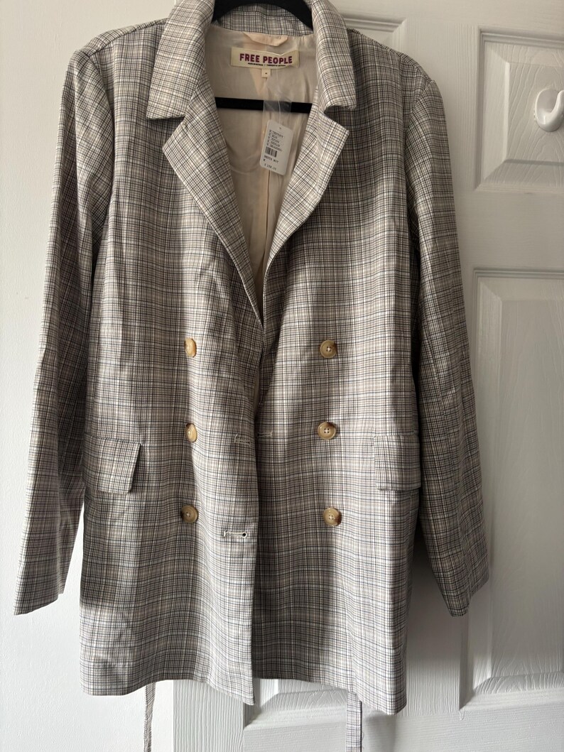 May include: A double-breasted blazer in a plaid pattern with tan, white, and gray tones. The blazer has a notched collar, two front pockets, and four buttons. The brand name "FREE PEOPLE" is visible on the inner label.