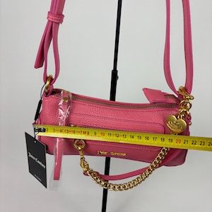 May include: A pink Juicy Couture shoulder bag with a gold chain strap and zipper. The bag is approximately 9 inches long. The bag has a gold heart charm and a tag that says Juicy Couture.