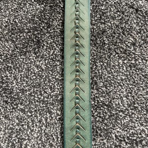 May include: Close-up of a teal leather belt with a decorative stitched pattern. The belt features a series of evenly spaced, arrow-shaped stitches running down the center. The leather has a slightly distressed appearance.