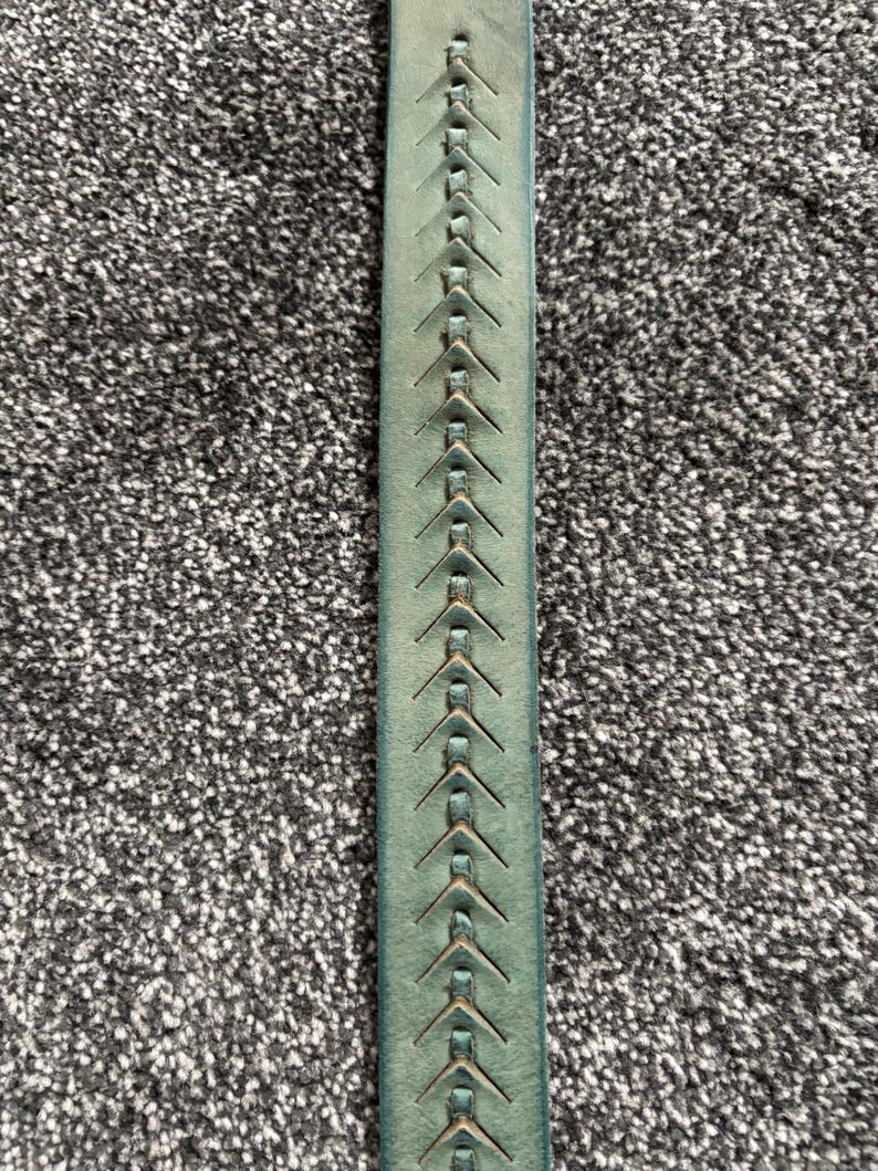 May include: Close-up of a teal leather belt with a decorative stitched pattern. The belt features a series of evenly spaced, arrow-shaped stitches running down the center. The leather has a slightly distressed appearance.