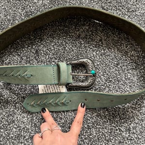 May include: A light green leather belt with a decorative silver buckle and turquoise accent. The belt features stitched detailing and a classic western design. The belt is shown on a gray surface.