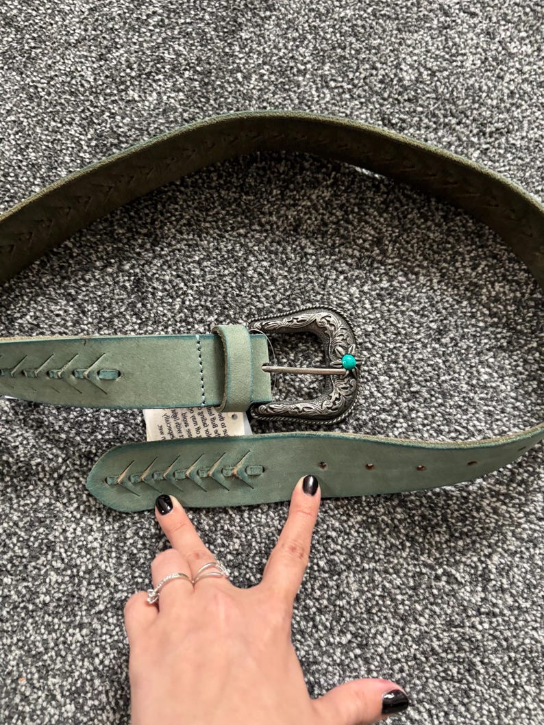 May include: A light green leather belt with a decorative silver buckle and turquoise accent. The belt features stitched detailing and a classic western design. The belt is shown on a gray surface.