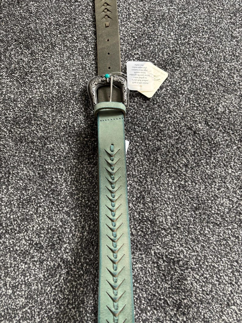 May include: A distressed green leather belt with a decorative silver buckle and turquoise accent. The belt features a series of cut-out designs along its length. A tag is attached to the belt.