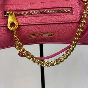 May include: A pink leather handbag with a gold-colored chain strap and zipper. The bag features the text "Juicy Couture" in gold lettering. The zipper pull and chain attachments are also gold-colored.