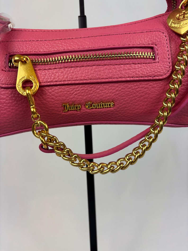May include: A pink leather handbag with a gold-colored chain strap and zipper. The bag features the text "Juicy Couture" in gold lettering. The zipper pull and chain attachments are also gold-colored.