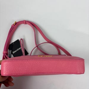 May include: A pink crossbody bag with a long strap and gold-colored hardware. The bag has a textured surface and a small tag attached. The brand name is visible in gold lettering on the bag.
