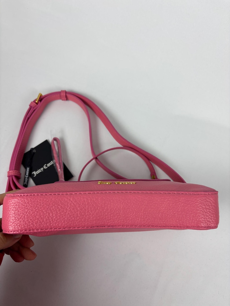 May include: A pink crossbody bag with a long strap and gold-colored hardware. The bag has a textured surface and a small tag attached. The brand name is visible in gold lettering on the bag.