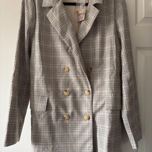 May include: A double-breasted blazer in a neutral plaid pattern. The jacket features large, round, light-colored buttons and two front pockets. The label reads "FREE PEOPLE". The blazer is hanging on a black hanger.