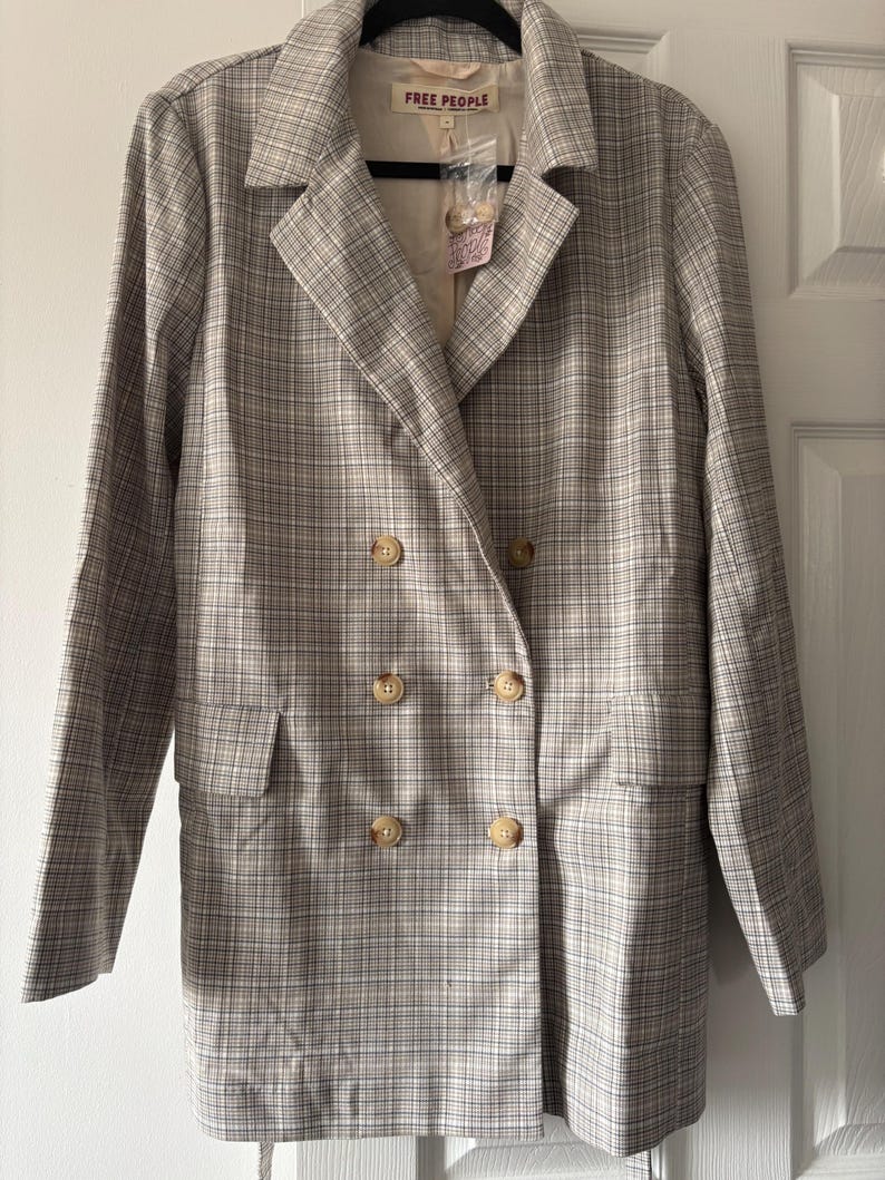 May include: A double-breasted blazer in a neutral plaid pattern. The jacket features large, round, light-colored buttons and two front pockets. The label reads "FREE PEOPLE". The blazer is hanging on a black hanger.