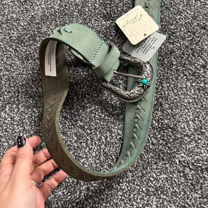 May include: A turquoise leather belt with a silver buckle and a turquoise stone accent. The belt has decorative stitching and a tag that reads "Free People". The belt is approximately 1.5 inches wide.