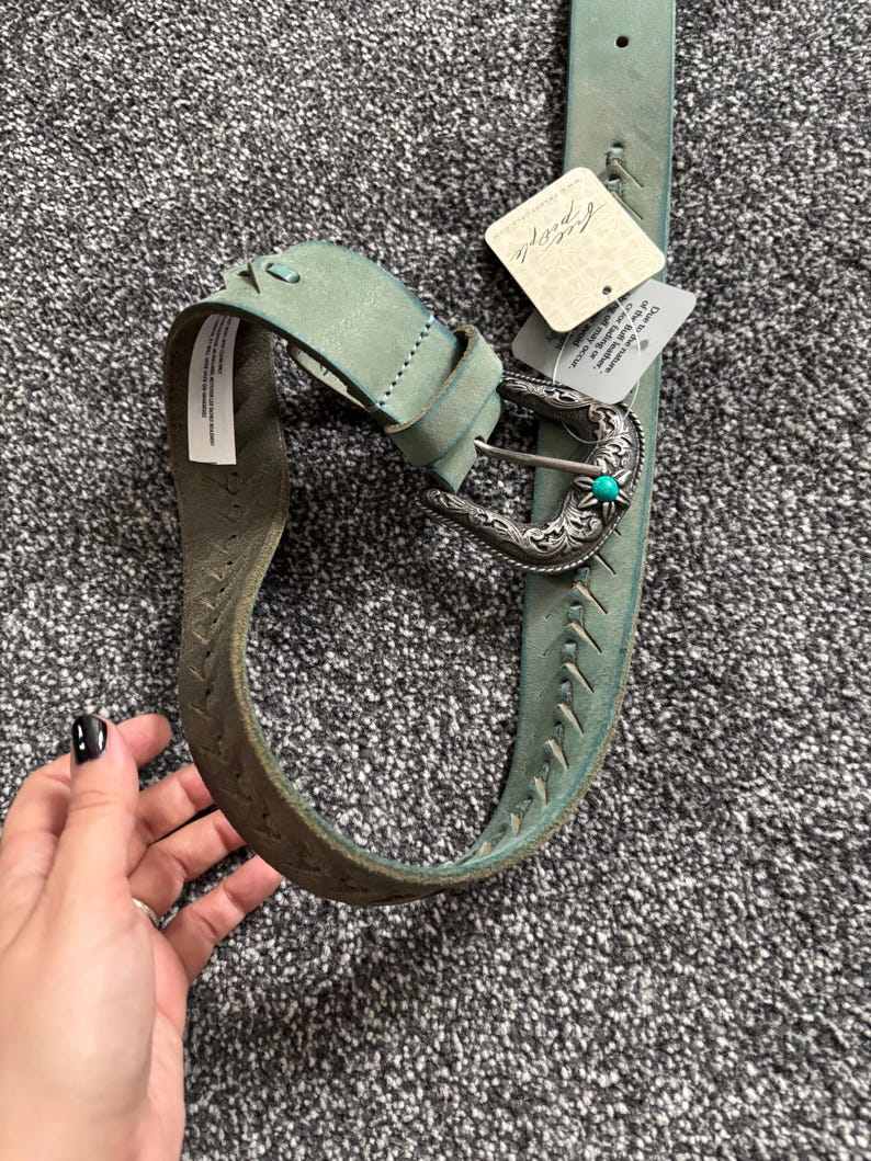 May include: A turquoise leather belt with a silver buckle and a turquoise stone accent. The belt has decorative stitching and a tag that reads "Free People". The belt is approximately 1.5 inches wide.