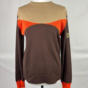 Puede incluir: Un suéter marrón de manga larga con una parte superior beige y un diseño de rayas naranjas. Los puños son naranjas y la manga izquierda tiene un logotipo dorado. El suéter se exhibe en un maniquí.