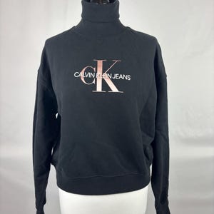 Op de afbeelding: Zwarte coltrui sweatshirt met het Calvin Klein Jeans logo in roségoud. De sweatshirt heeft lange mouwen en een geribde zoom. Het logo staat in het midden op de borst. Het kledingstuk wordt op een mannequin gepresenteerd.