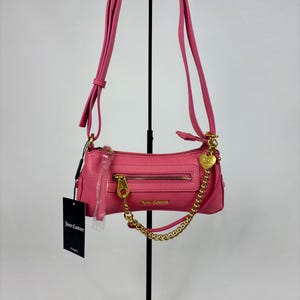 May include: A pink, rectangular Juicy Couture handbag with a gold zipper and chain strap. The bag has a long, adjustable strap and a small heart-shaped charm. A black tag with the Juicy Couture logo hangs from the bag.