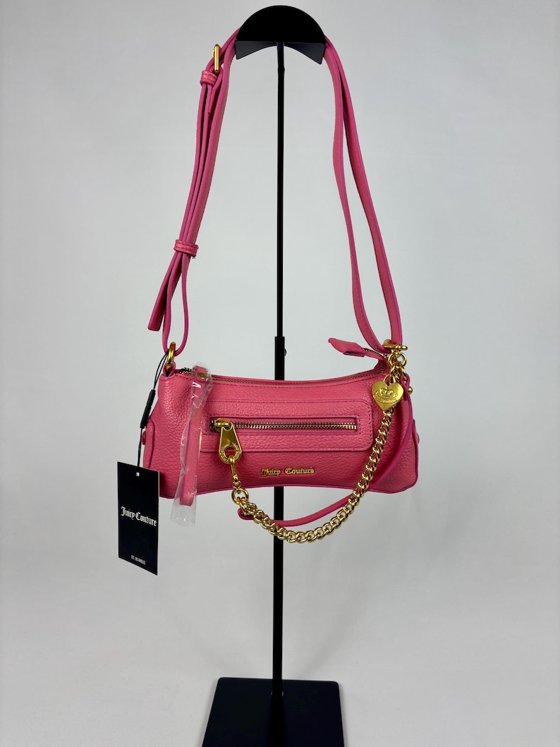 May include: A pink, rectangular Juicy Couture handbag with a gold zipper and chain strap. The bag has a long, adjustable strap and a small heart-shaped charm. A black tag with the Juicy Couture logo hangs from the bag.