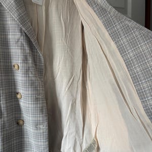 May include: A plaid blazer in shades of gray and white, featuring a double-breasted design with three visible buttons. The interior lining is a light beige color. A tag with text is attached to the blazer.