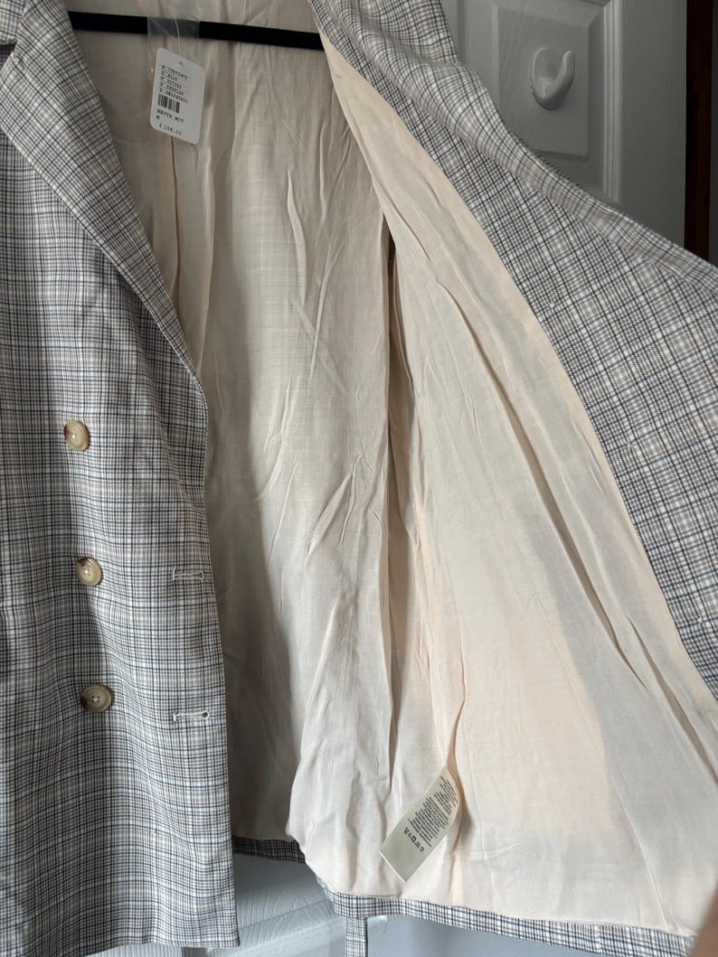 May include: A plaid blazer in shades of gray and white, featuring a double-breasted design with three visible buttons. The interior lining is a light beige color. A tag with text is attached to the blazer.