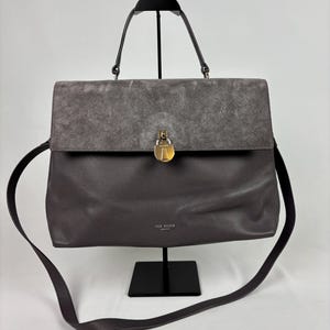 May include: A grey leather handbag with a suede flap and a gold-tone lock. The bag features a top handle and a long shoulder strap. The brand name "TED BAKER" is visible on the front of the bag.