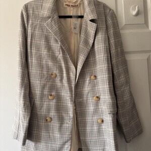 May include: A tan and white plaid blazer with a double-breasted design. The blazer has four buttons and a notched lapel. The brand "FREE PEOPLE" is visible on the label. The blazer is hanging on a black hanger.