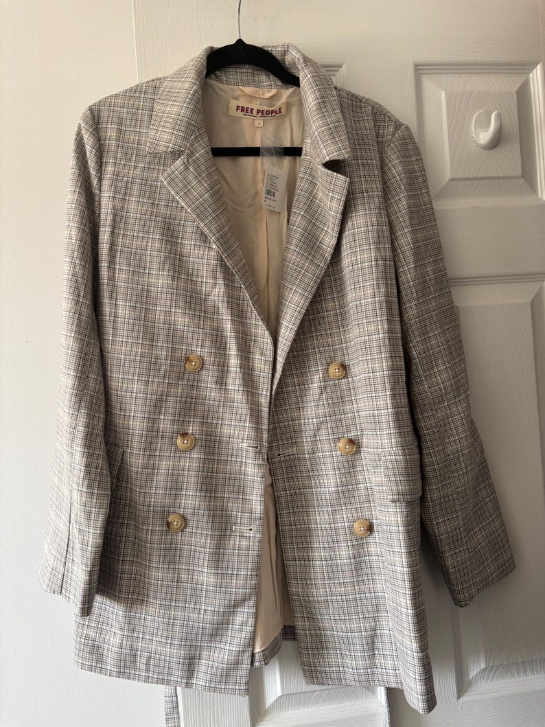 May include: A tan and white plaid blazer with a double-breasted design. The blazer has four buttons and a notched lapel. The brand "FREE PEOPLE" is visible on the label. The blazer is hanging on a black hanger.