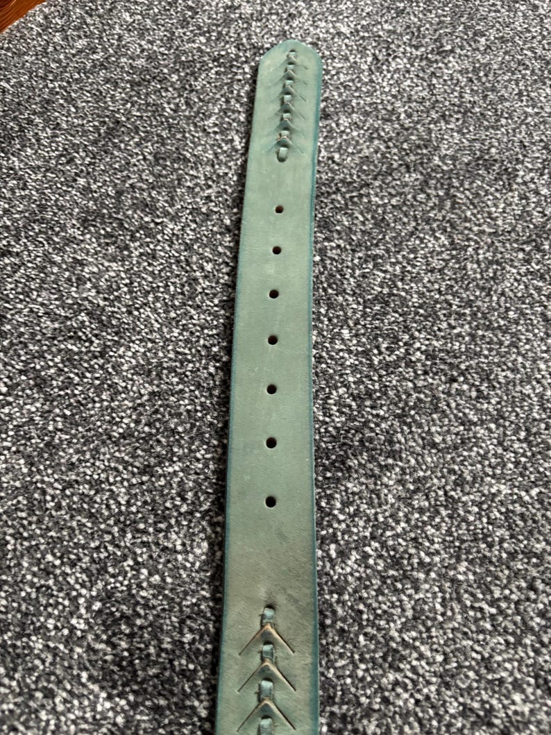 May include: A teal-colored leather belt with decorative stitching and multiple holes for adjustment. The belt has a slightly distressed appearance, suggesting it is made of genuine leather. The design includes arrow-shaped details near the buckle.