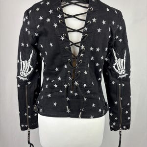 May include: Black denim jacket with white star pattern and lace-up back. Features zipper sleeves and embroidered skeleton hand patches. A unique, edgy piece of outerwear.